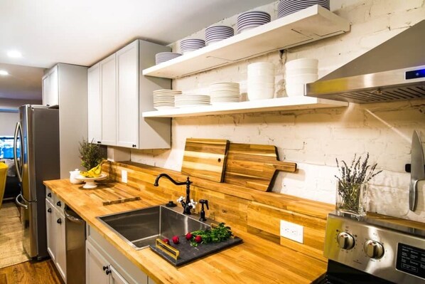 A kitchen with a wooden countertop, stainless steel sink, and a built-in stove. There are white cabinets and a shelf with plates above the counter. - Yellow Block BnB (Brooklyn)