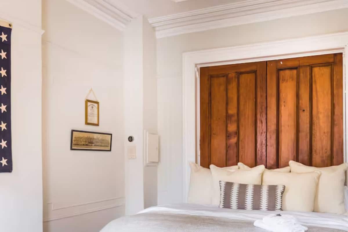 A neatly made bed with a wooden headboard, a bedside table with books, and a small vase.