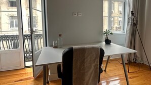 Une pièce avec un bureau blanc, un fauteuil de bureau noir sur lequel est posée une serviette, une plante en pot et une vue sur des immeubles à travers de grandes fenêtres.