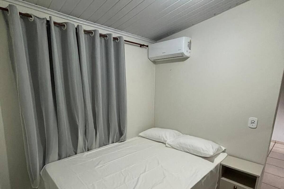 Une chambre avec un lit simple, un couvre-lit blanc, des rideaux gris, un appareil de climatisation, une table de chevet et un sol carrelé.