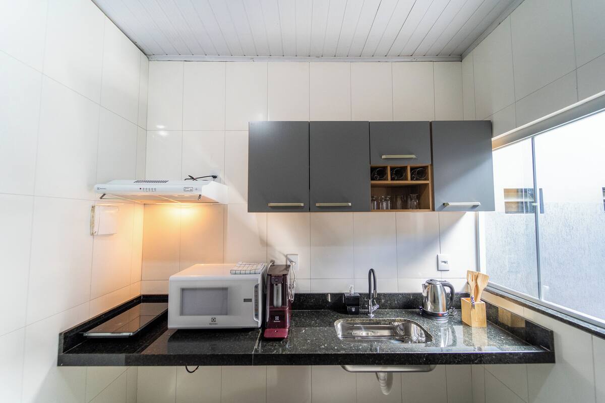 Une cuisine compacte avec des murs recouverts de carreaux blancs, un plan de travail noir, un four à micro-ondes, une machine à café, un évier et une fenêtre qui laisse entrer la lumière naturelle.
