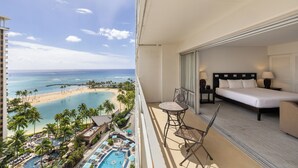 A balcony with a bed, a chair, and a view of a beach and pool area. - Sweeping Ocean Views | Ilikai Hotel Waikiki 1534 – Heated Pool & Spa (Honolulu)