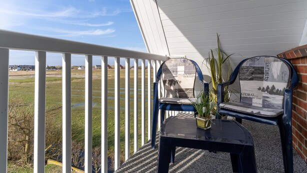 A balcony with a view of a grassy field and a brick wall. There are two chairs with a plant on a table. - Ferienwohnung fur 4 in St. Peter-ording (St. Peter-Ording)