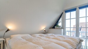 A bedroom with a large bed, two bedside lamps, a window with blue curtains, and a view of buildings outside. - Ferienwohnung fur 4 in St. Peter-ording (St. Peter-Ording)