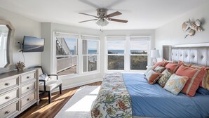Un dormitorio con cama, cómoda, televisor y ventanas amplias con vista al océano.