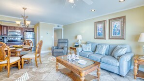 A spacious living area with a dining table, chairs, a sofa, and a coffee table. There is a ceiling fan, a kitchen in the background, and two framed pictures on the wall.