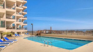 A swimming pool with blue lounge chairs, a concrete deck, and a building with balconies.