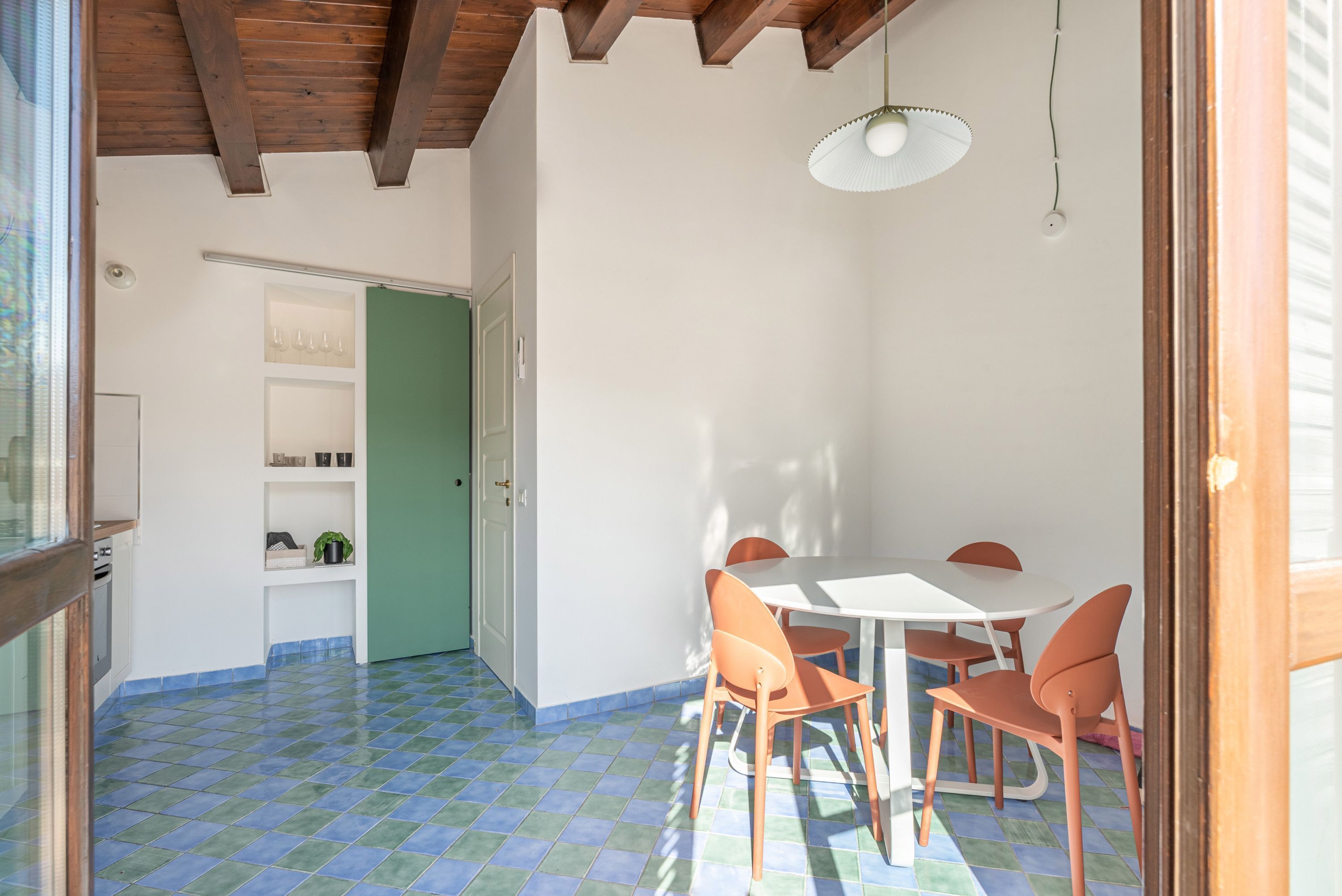 Zona pranzo con tavolo bianco e sedie arancioni, una porta verde e pavimento piastrellato.
