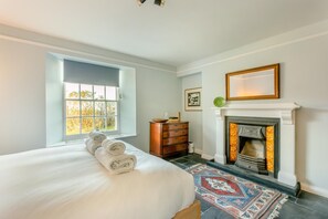 A bedroom with a large bed, a fireplace, a dresser, and a window with a view of trees.