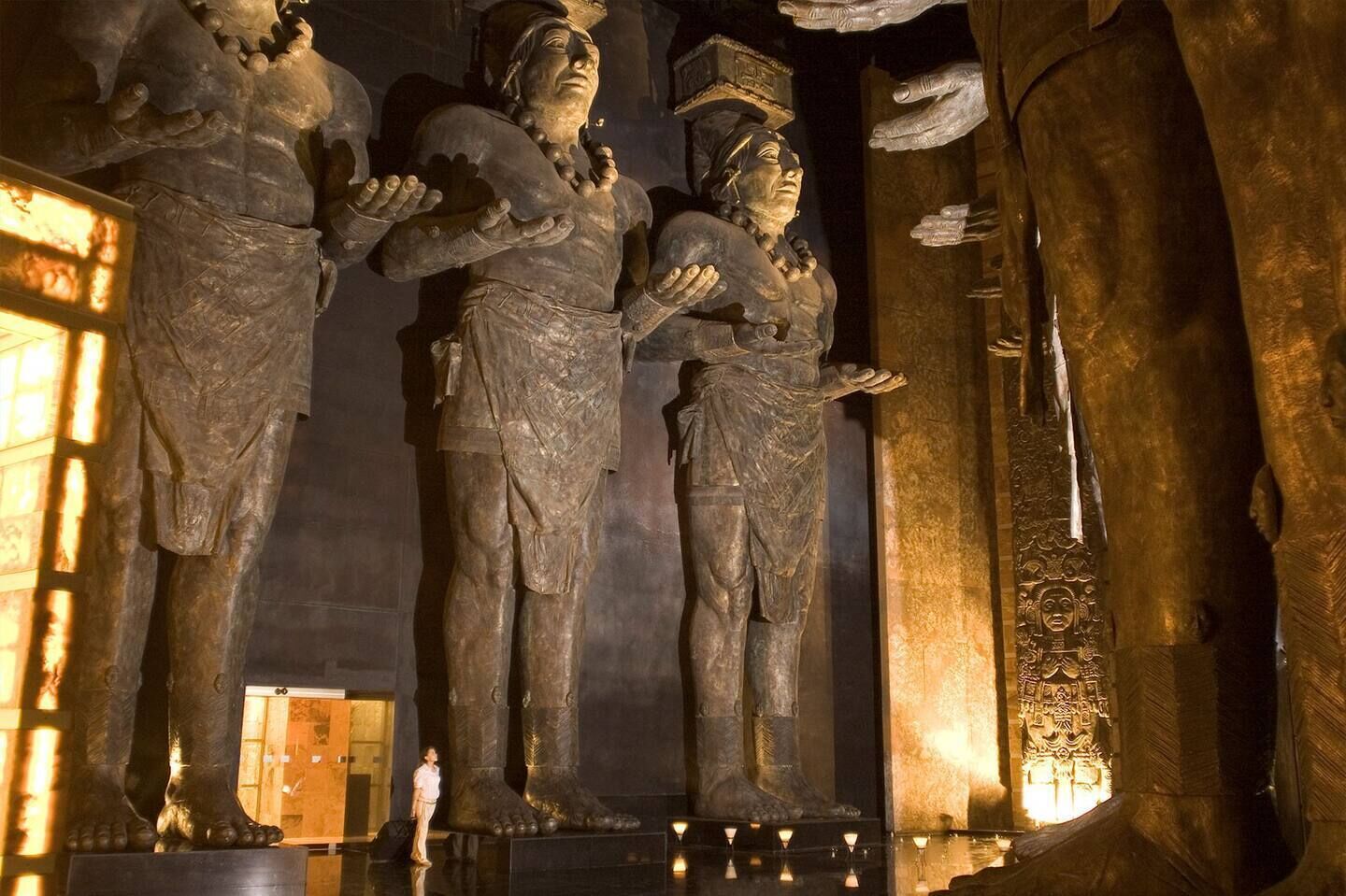 A large, ancient stone statue with intricate carvings, standing in a dimly lit room with a reflective floor.