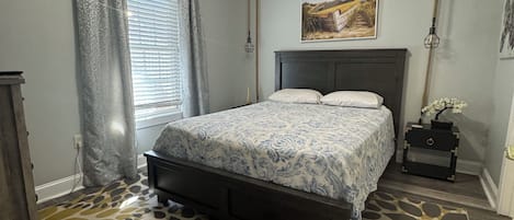 A bedroom with a bed, a ceiling fan, a window with curtains, a nightstand with a lamp, and a patterned rug.