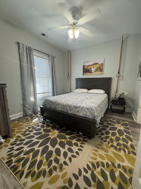 A bedroom with a bed, a ceiling fan, a window with curtains, a nightstand with a lamp, and a patterned rug.