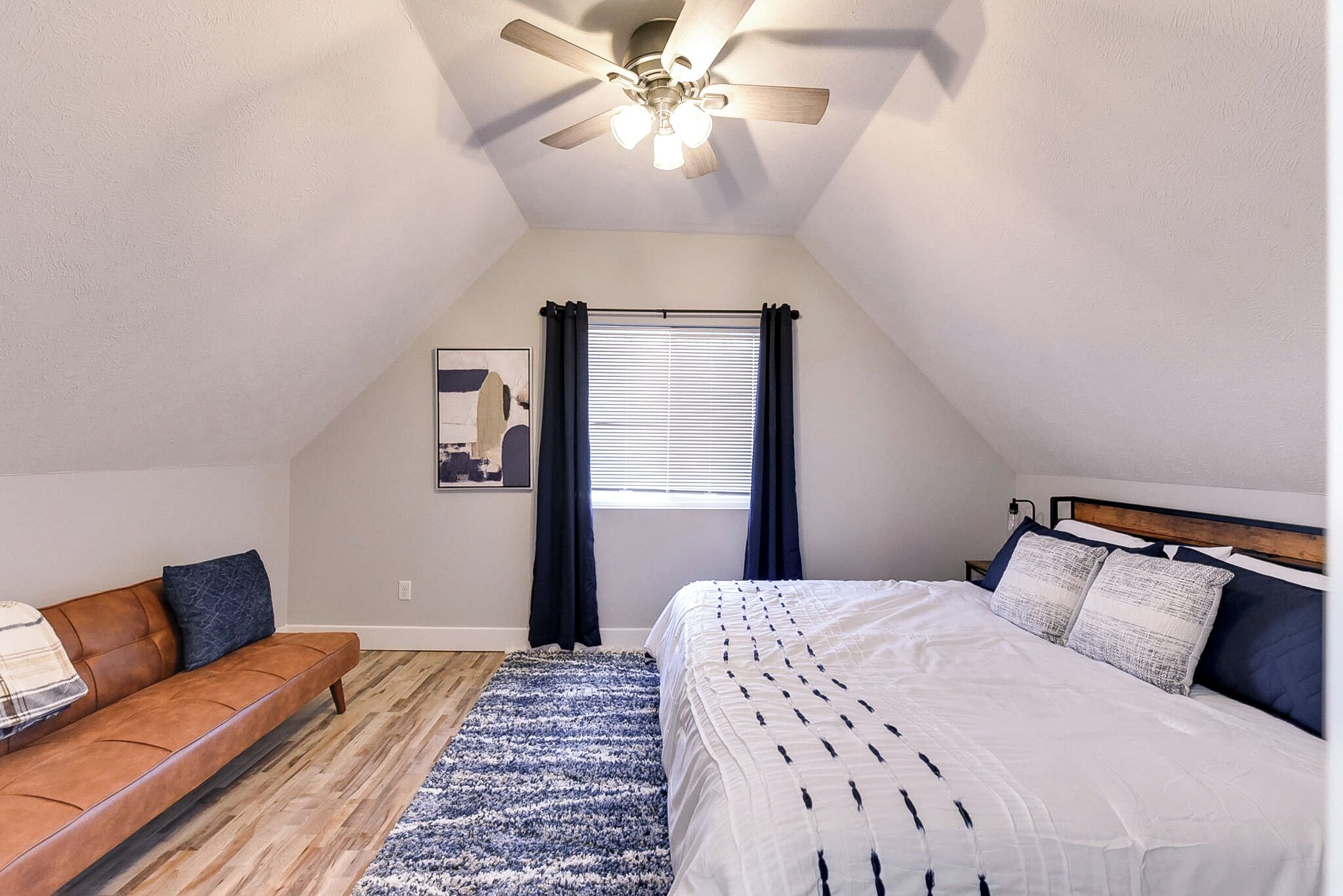 Une chambre à coucher comprenant un lit, un canapé, un ventilateur de plafond, une fenêtre avec des rideaux et une œuvre d’art accrochée au mur.