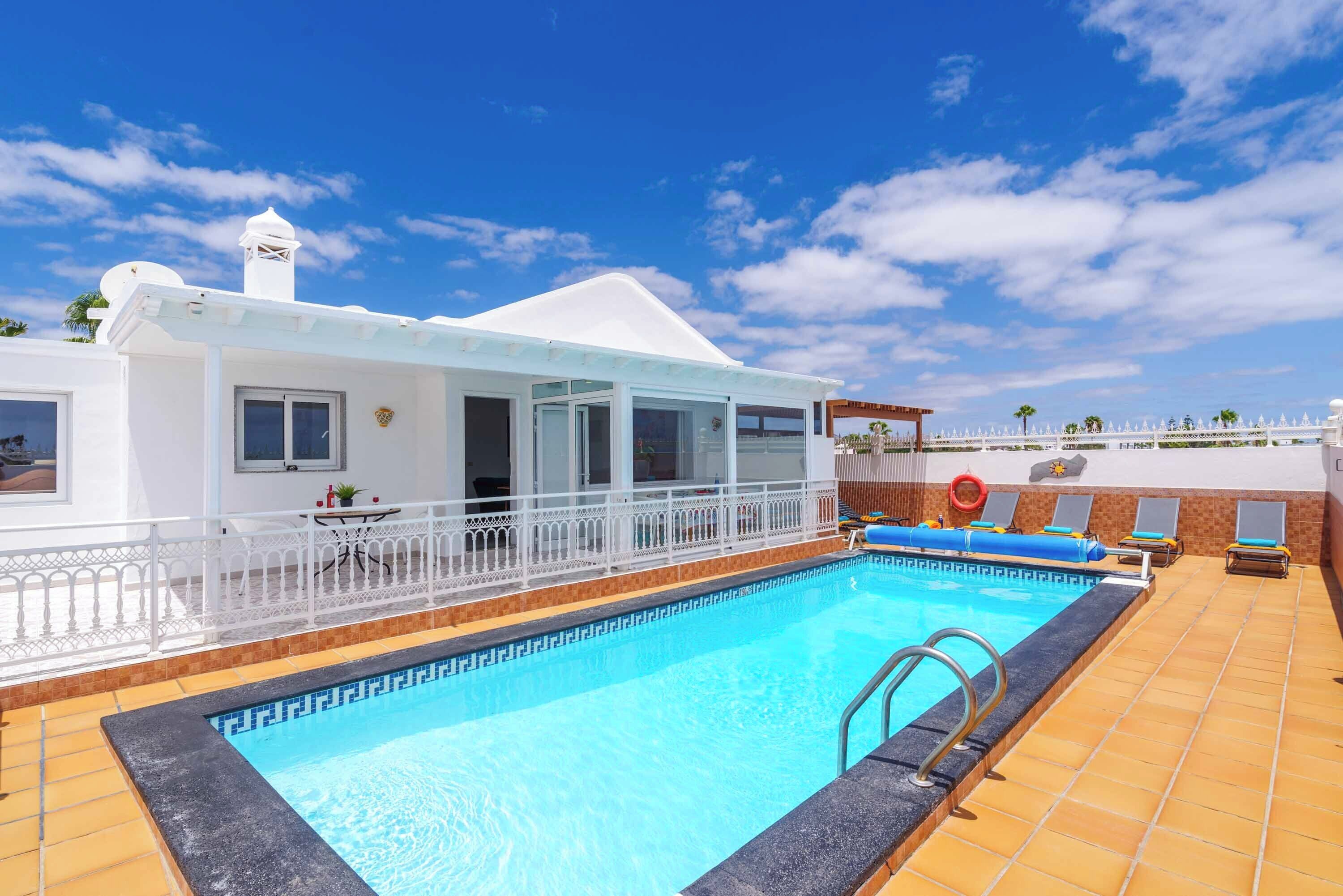 Une piscine avec un bâtiment blanc, un espace abrité et une clôture blanche.