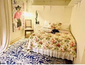 Une chambre à coucher avec une couvre-lit à motifs floraux, une petite table de chevet avec une lampe et une affiche accrochée au mur.