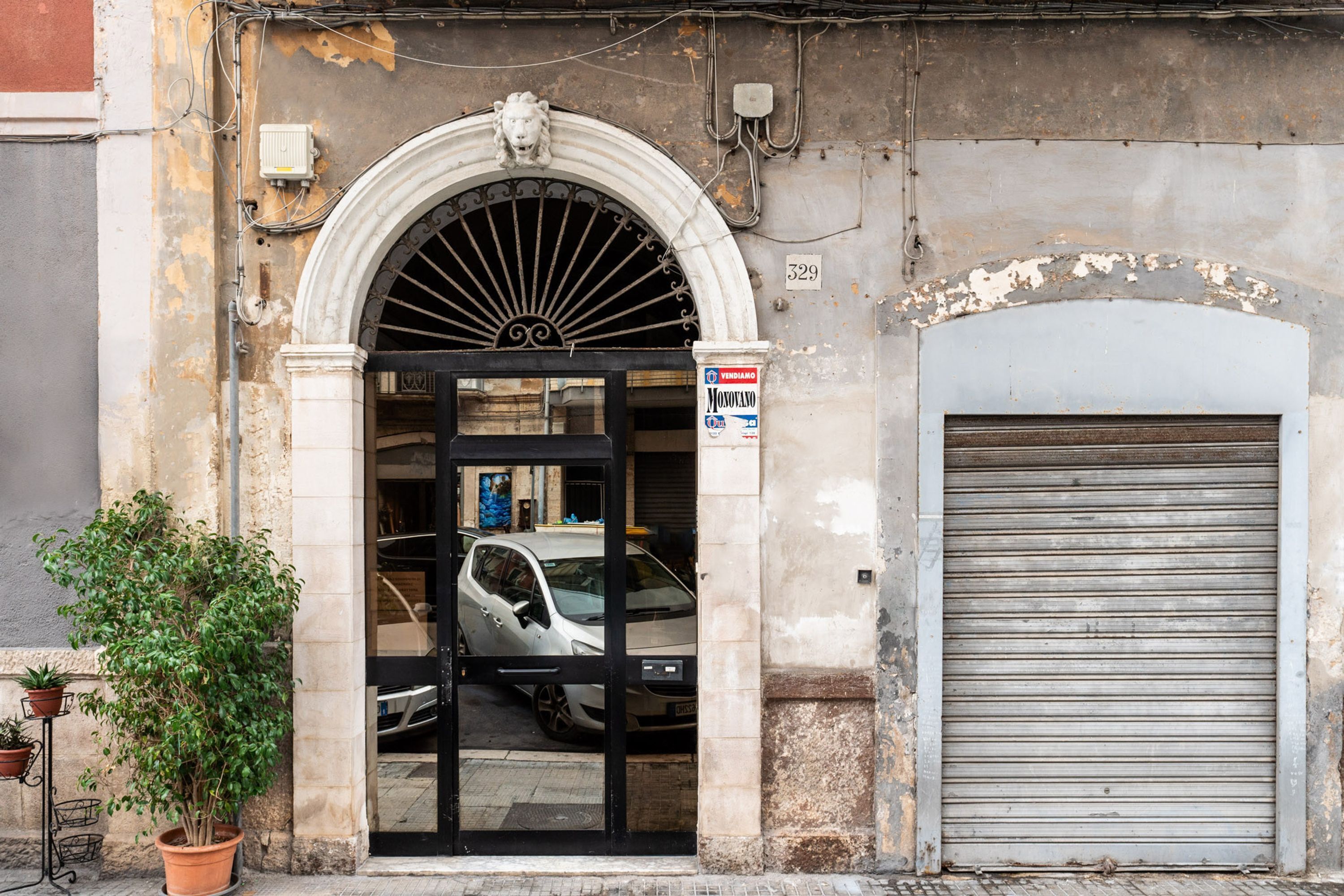 L'ingresso di un edificio con un cancello nero, un'auto in un box chiuso e una pianta in vaso.