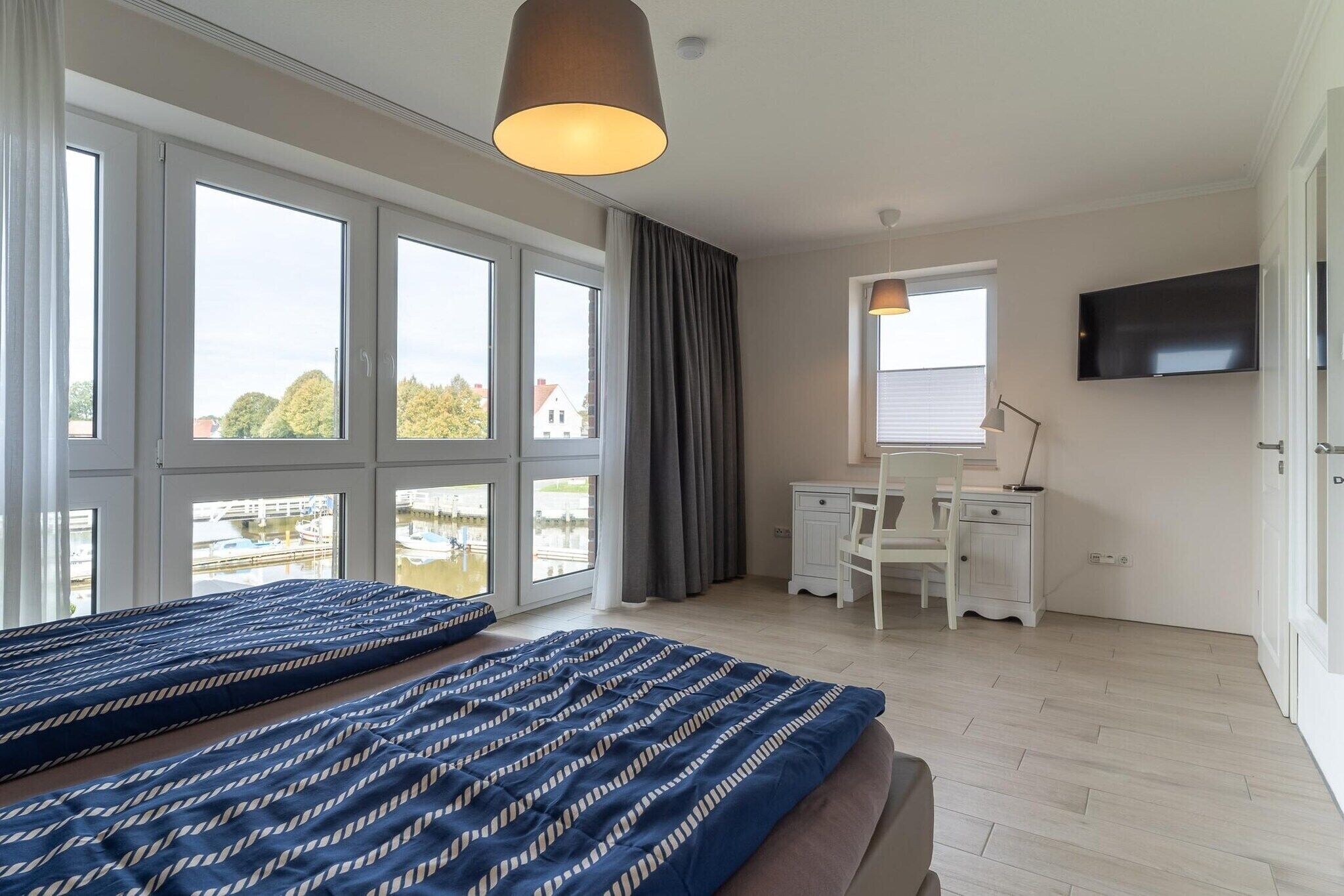 A bedroom with a bed, a desk, a chair, a TV, and large windows.
