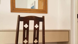 Een houten stoel met een beklede zitting en rugleuning, geplaatst voor een witte muur met daarboven een spiegel met houten lijst.