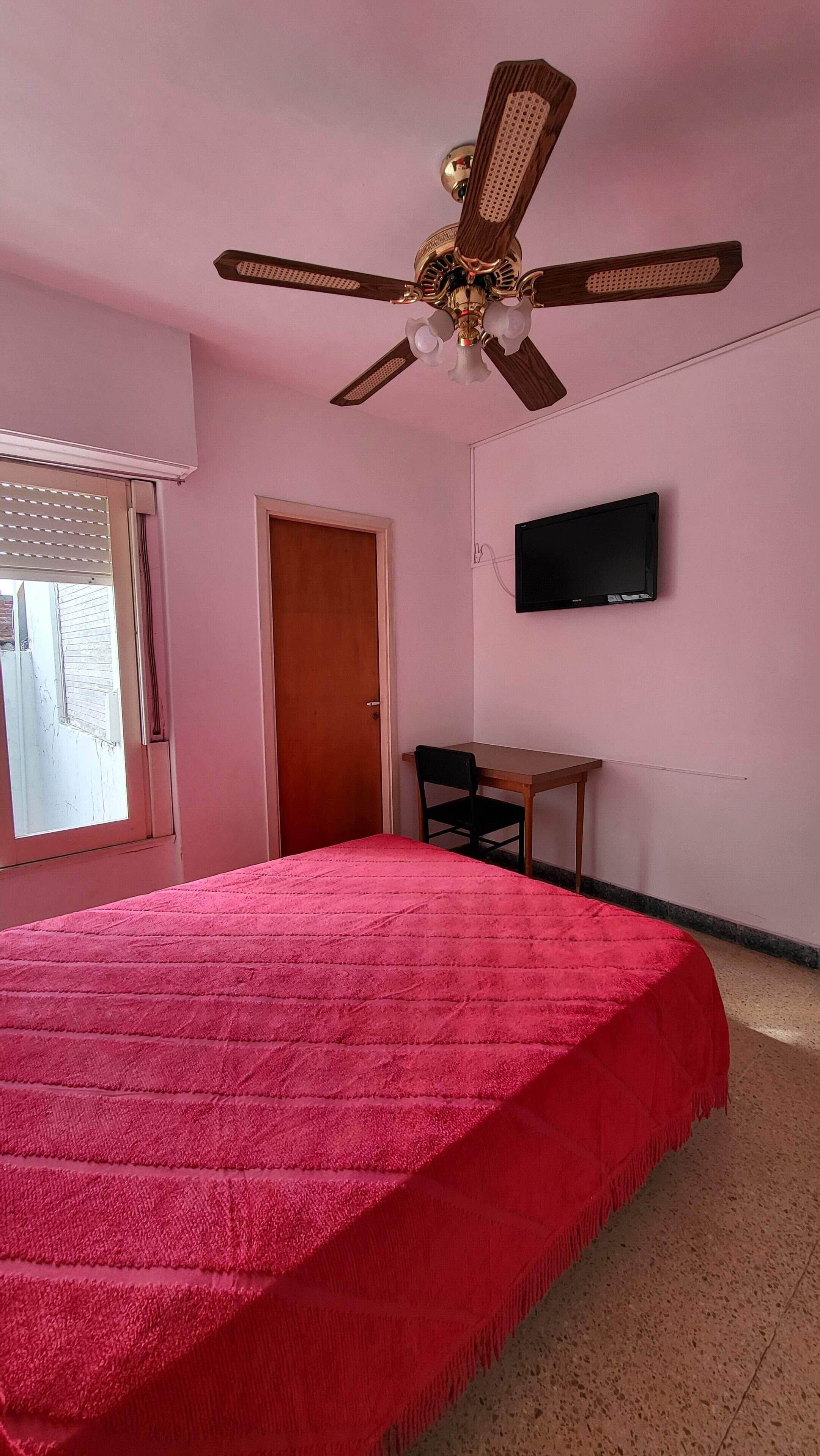 A bedroom with a bed, a ceiling fan, a TV, and a window with blinds.