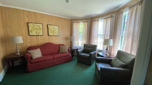 Una sala de estar con un sofá rojo, dos sillones y una mesa de centro. Hay dos lámparas sobre mesitas auxiliares y el cuarto tiene paneles de madera en las paredes.
