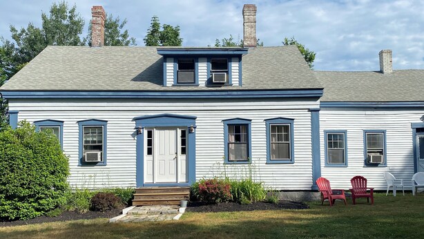 Une maison à deux étages avec un porche à l’avant, une cheminée et un petit jardin agrémenté de chaises.