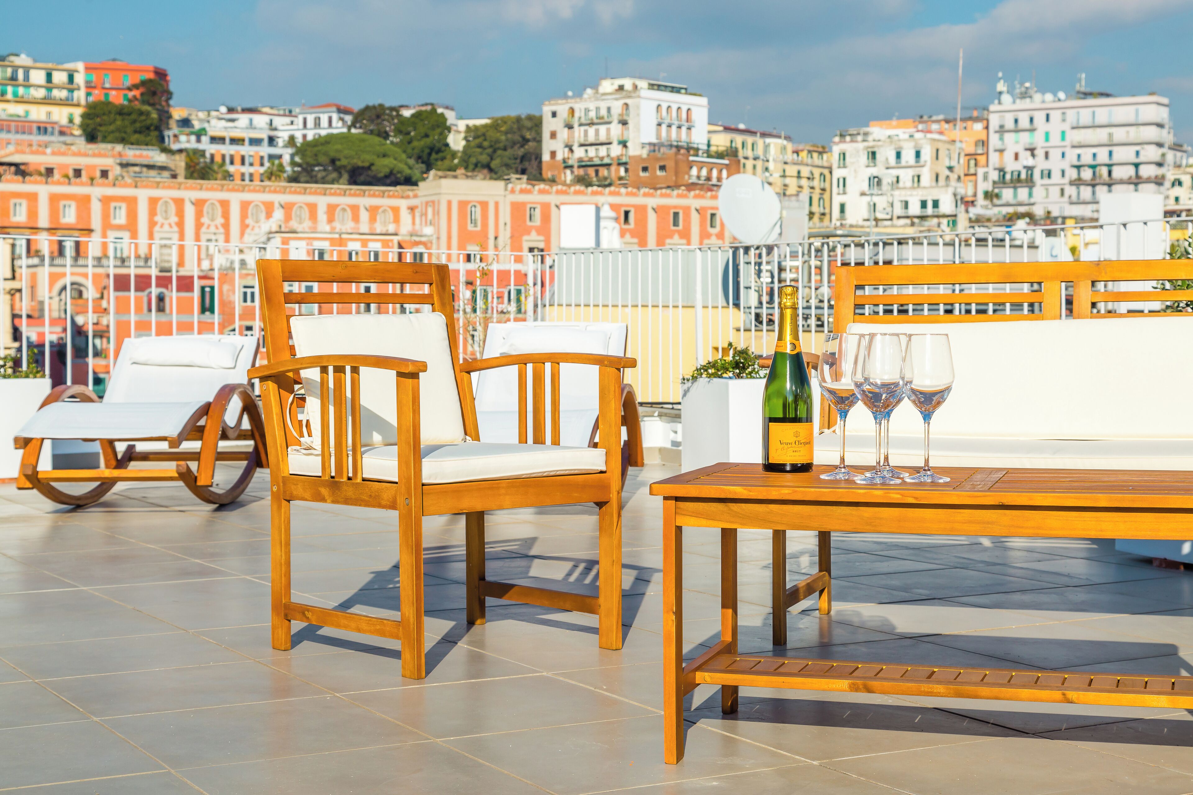 Una terraza con mobiliario de madera, una mesa con una botella y vasos, y vista a un paisaje urbano.