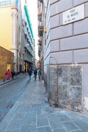 Una calle adoquinada con edificios históricos, un cartel que indica Piazza de Marti, y peatones.