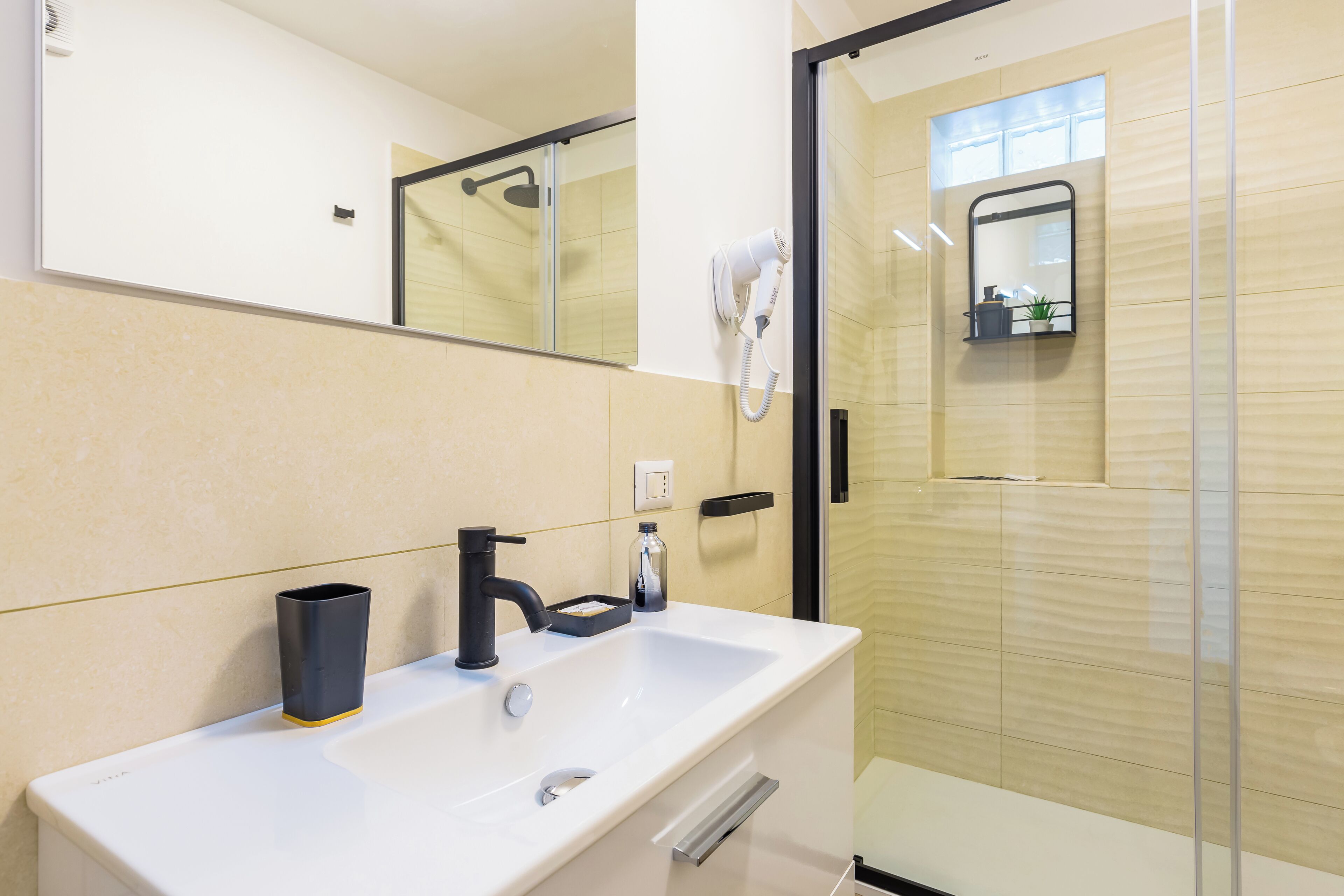 A bathroom with a white sink, black faucet, and a mirror above it. There is a towel hanging on the wall and a small shelf with toiletries.