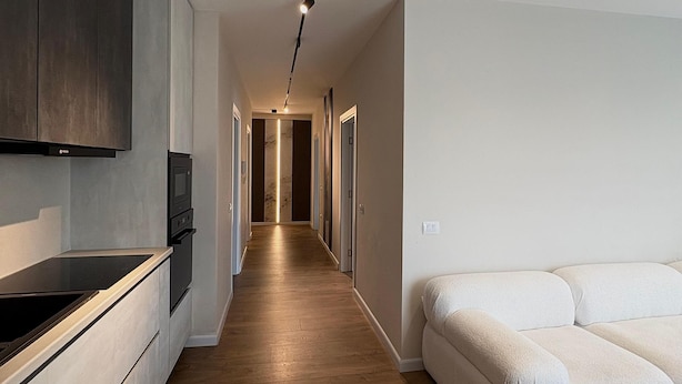 Eine moderne Küche mit dunkler Holz Arbeitsplatte, einem eingebauten Backofen und einer Mikrowelle. Ein langer Flur führt zu einem Schlafzimmer mit einem weißen Sofa.