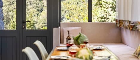 A dining area with a table set for two, a sofa, and a view of a forested landscape through large windows.