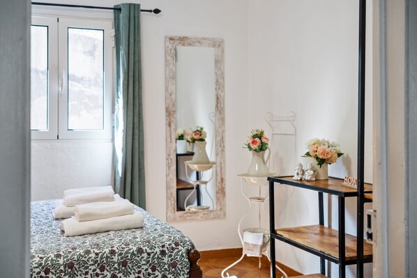 A neatly made bed with folded towels, a bedside table with a vase of flowers, a mirror on the wall, and a window with curtains.