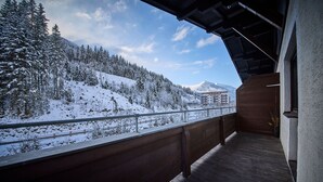 Un balcon doté d’un plancher et de balustrades en bois, offrant une vue sur un paysage enneigé, avec des arbres et une montagne en arrière-plan.