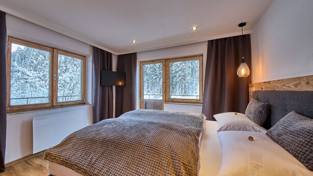 Une chambre à coucher avec un grand lit, deux fenêtres avec des rideaux, un téléviseur fixé au mur et une tête de lit en bois.