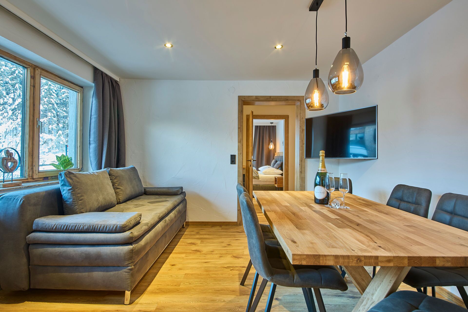 A modern living room with a wooden dining table, sofa, and a TV mounted on the wall.