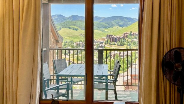 Ein Balkon mit Tisch, Stühlen, einem Ventilator und Blick auf Berge und Gebäude.