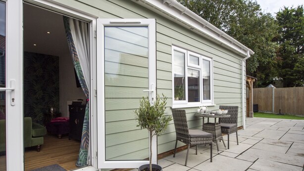 Une maison dotée d’une porte-fenêtre coulissante donnant sur un patio aménagé avec une table et des chaises, un pot de fleurs et une terrasse en bois.