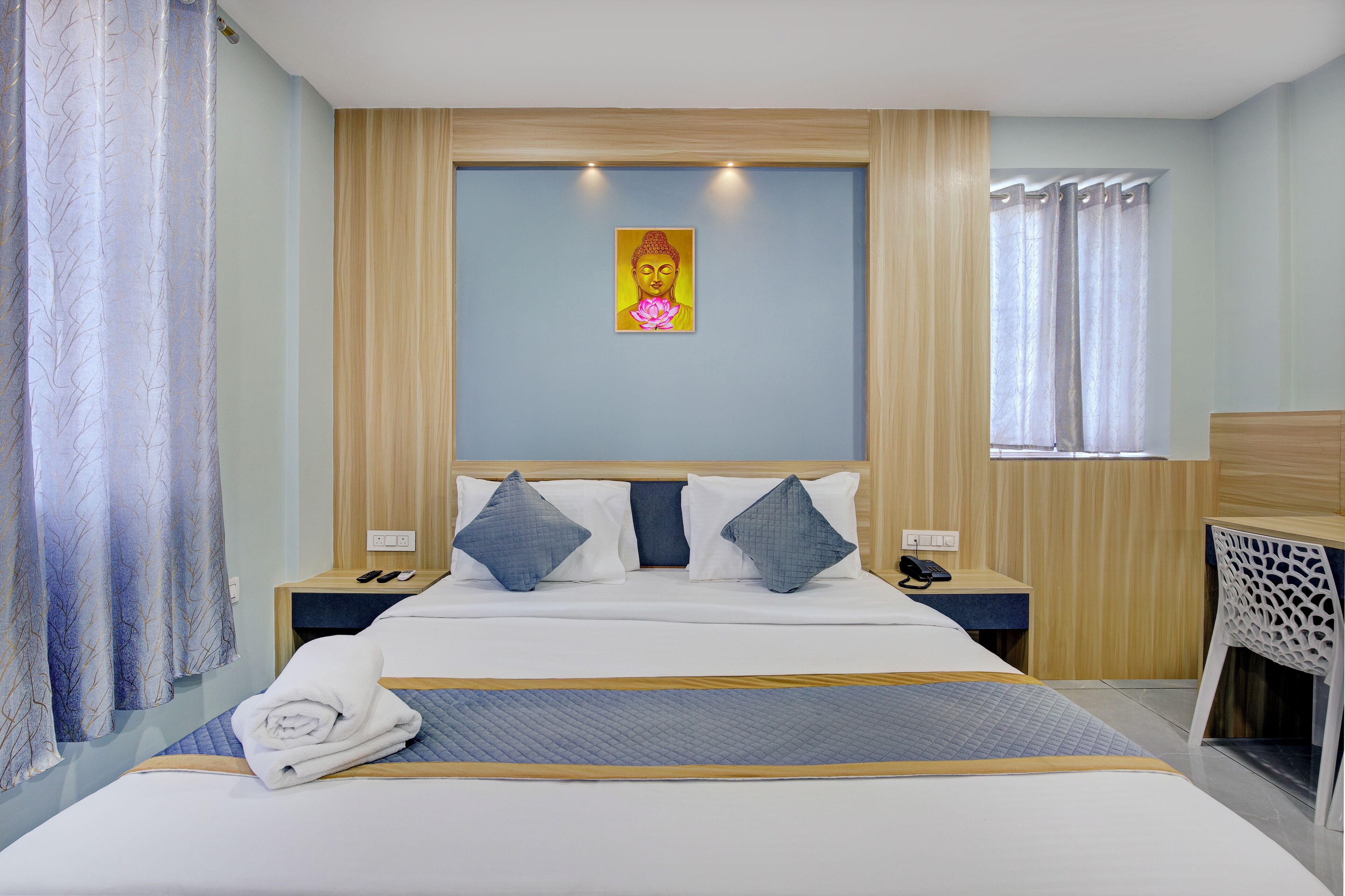 A hotel room with a bed, two bedside tables, a nightstand, a window with curtains, and a wall-mounted picture.