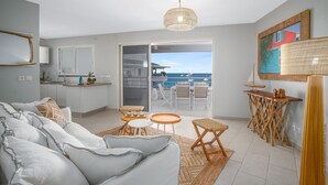 A modern living room with a sofa, coffee table, and chairs, a kitchen area, and a balcony with a view of the sea. - Horizon Bleu (Trois-Ilets)