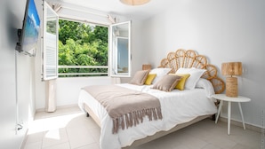 A bedroom with a bed, a TV, a window with an open shutter, and a view of greenery outside. - Horizon Bleu (Trois-Ilets)