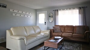 Een woonkamer met een beige bank, een bruine leren bank, een bijzettafeltje en een raam met gordijnen.