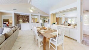 A spacious living area with a wooden dining table set for four, surrounded by white chairs. A sofa and a kitchen area are visible in the background.