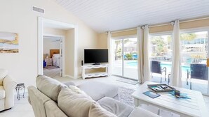 A living room with a sofa, a coffee table, and a TV. There is a door leading to a balcony with a pool view.