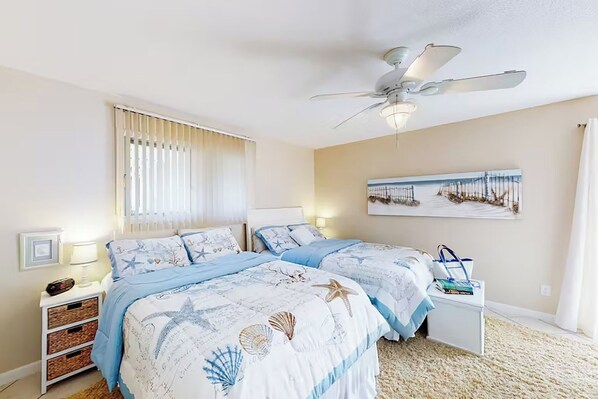 Une chambre avec deux lits, un ventilateur de plafond, une table de chevet, une lampe et un tableau encadré au mur.