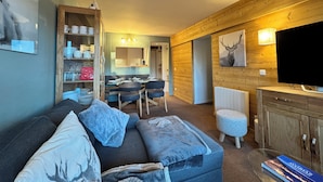 Ein gemütliches Wohnzimmer mit einer Couch, Hocker und einem Fernseher. Es gibt eine Essecke mit Stühlen und einem Tisch, und im Hintergrund ist eine Küche zu sehen.