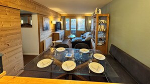 Ein gemütliches Blockhaus-Interieur mit Essbereich, inklusive Glastisch für vier Personen, Holzwänden, einer Couch und einem Schrank mit Gläsern.