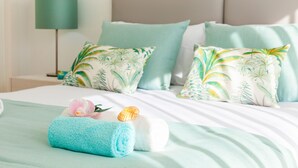 A neatly made bed with a teal bedspread, two pillows with a tropical leaf pattern, and a rolled towel with a pink flower and seashell. - Villa Aqua 4 (Albufeira)