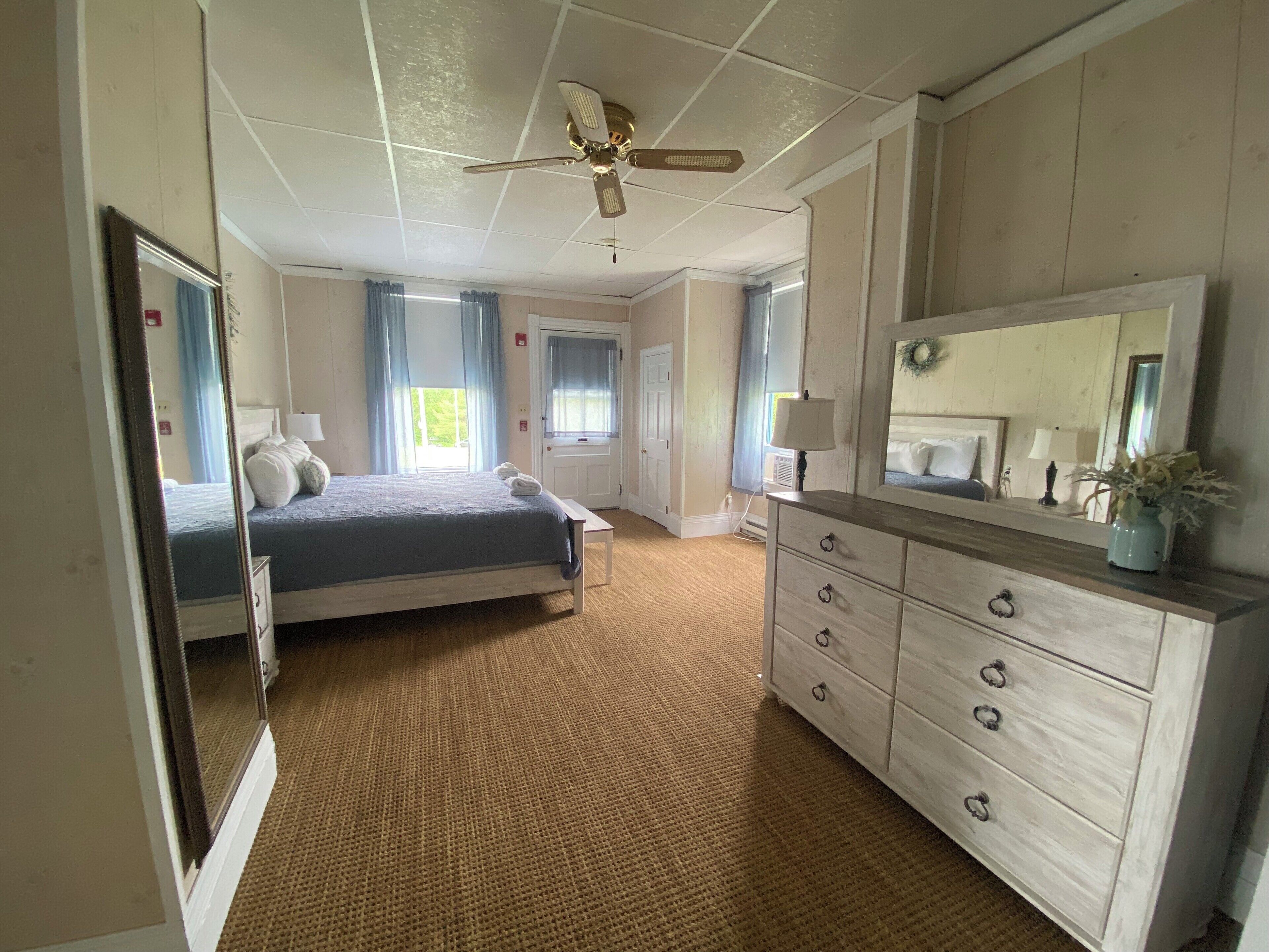 Quarto de hotel com cama, penteadeira, ventoinha no teto e janela com cortinas.