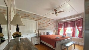 Ein Schlafzimmer mit Holzboden, einem Bett mit roter Decke, einem Nachttisch mit Lampe, einem Deckenventilator und einem Fenster mit roten Vorhängen.