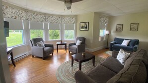 Ein Wohnzimmer mit Deckenventilator, zwei Sesseln, einer Couch, einem Couchtisch und einem Teppich. Es gibt Fenster mit Vorhängen und zwei gerahmte Bilder an der Wand.