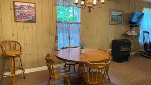 Un comedor con una mesa redonda de madera, cuatro sillas de madera, una araña, un ventanal con cortinas estampadas y un televisor sobre un mueble.